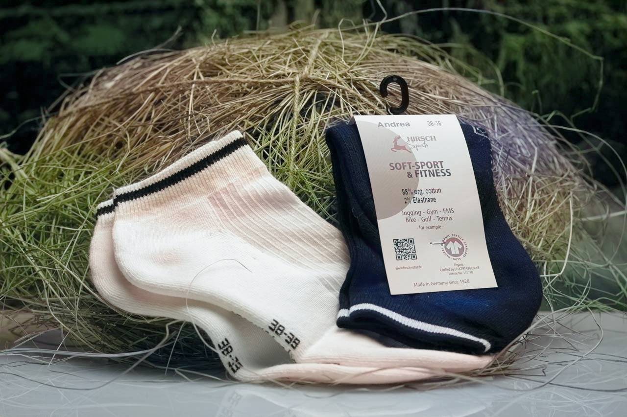 Ein Paar weiße und ein Paar marineblaue Sportsocken von 'Hirsch Natur Soft-Sport & Fitness' liegen vor einem Heuhintergrund.